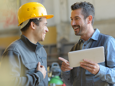 Smiling man holding a tablet talking to another smiling man wearing a hard hat in a warehouse.